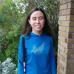 Picture of Áine, a white non-binary person with long dark hair, half-smiling, holding a white cane and wearing a blue hoodie.