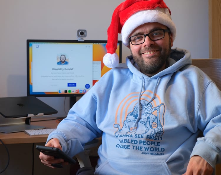 Picture of Peter at his desk, smiling with a Santa hat and Judy Heumann hoodie.
